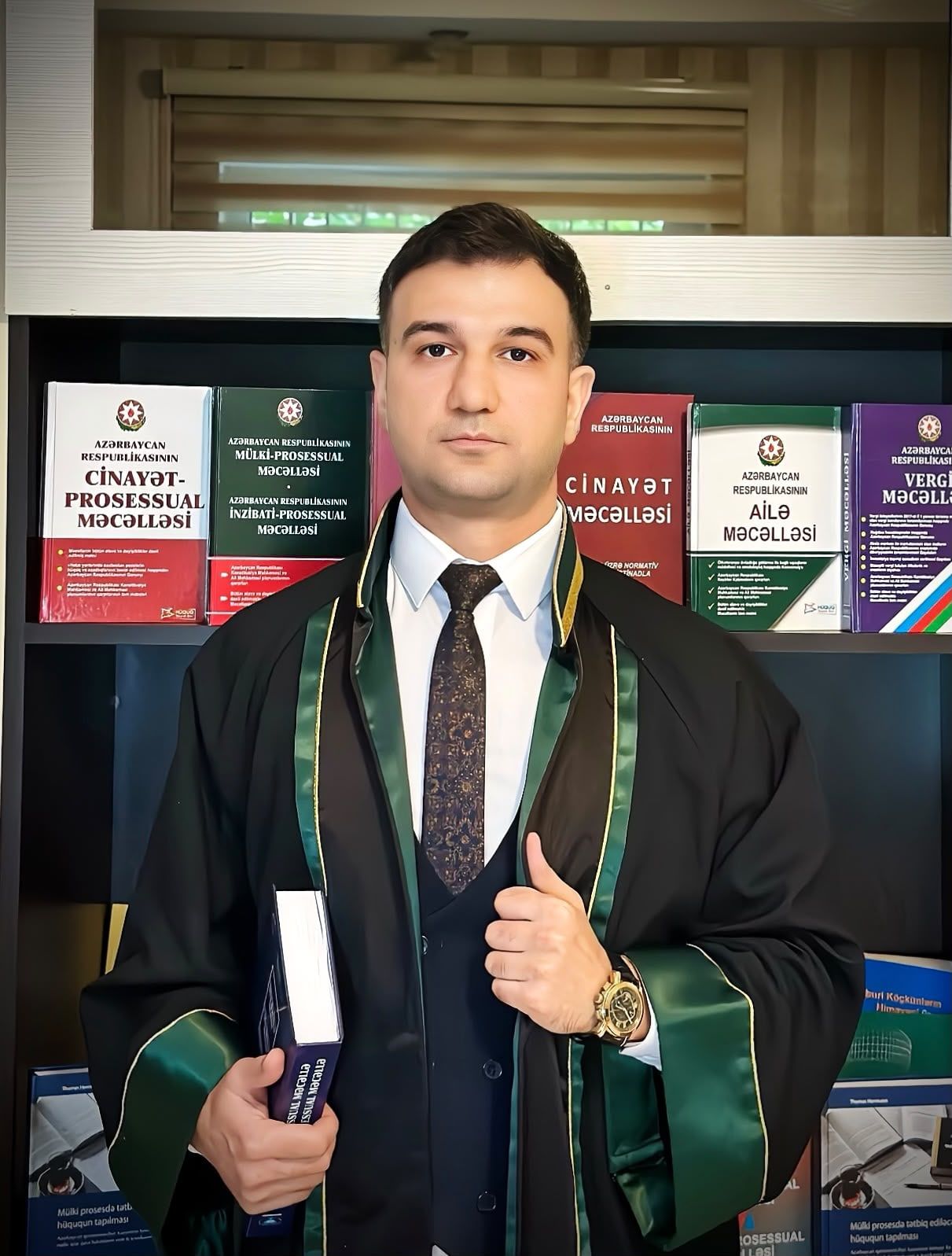 Vəkil Amid Əsgərov mantiyada, qanun məcəllələri olan rəfin qarşısında